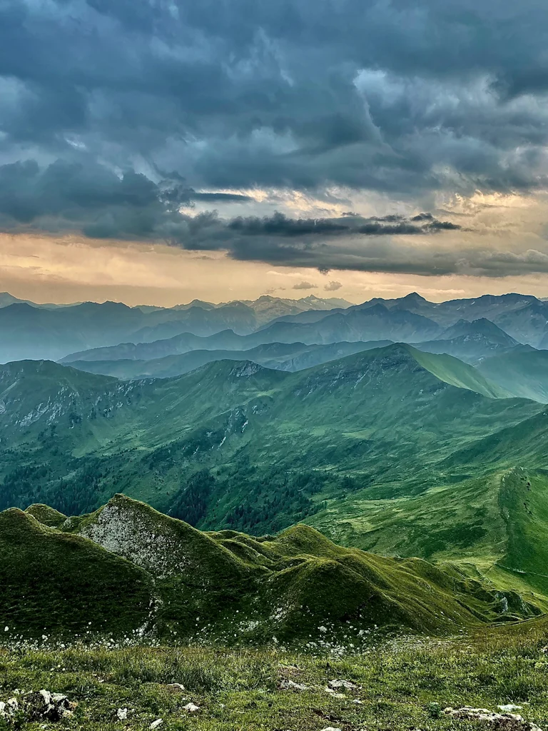 A panorama showing the peaks of the Austrian Alps at twilight. The sun is going down and green waves of mountain peaks are sitting beneath a gold and blue sky.
